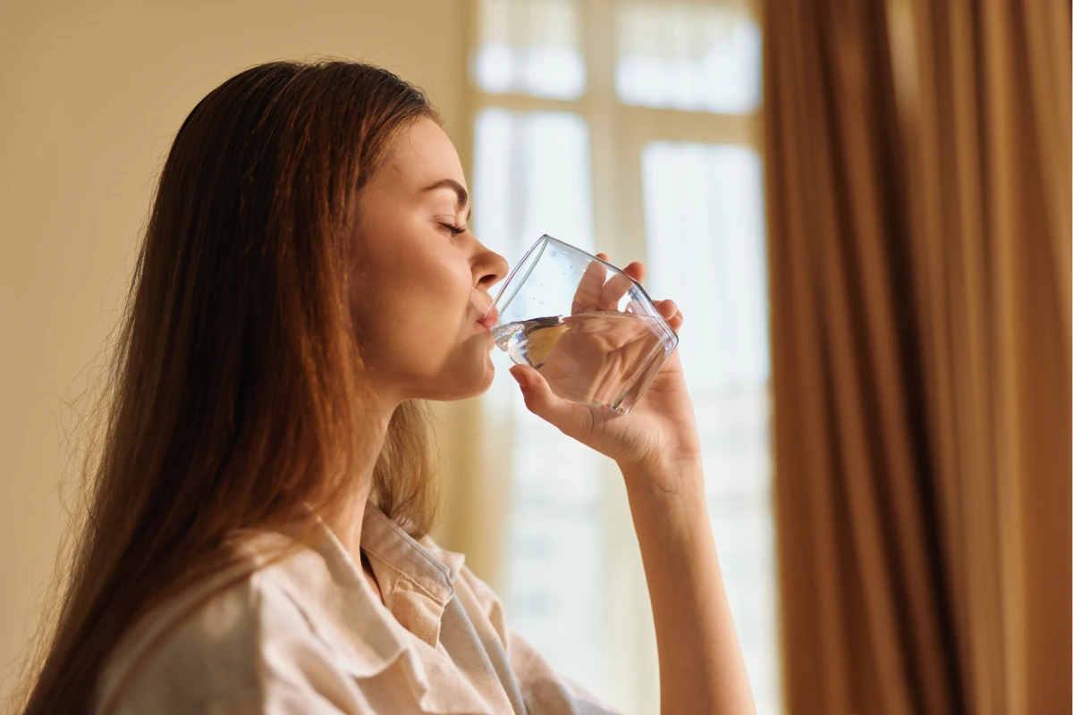Mujer que bebe el agua del vidrio en casa, vista lateral del retrato de la mujer joven relajada que hidrata adentro cerca de la ventana y de las cortinas, rutina de la mañana y estilo de vida sano.