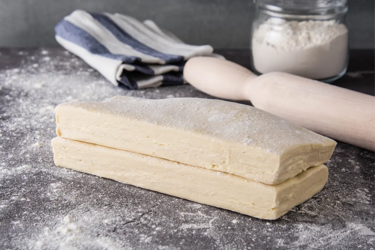 Dos masas de hojaldre apiladas que la acompaña un rodillo de madera y un bote con harina.