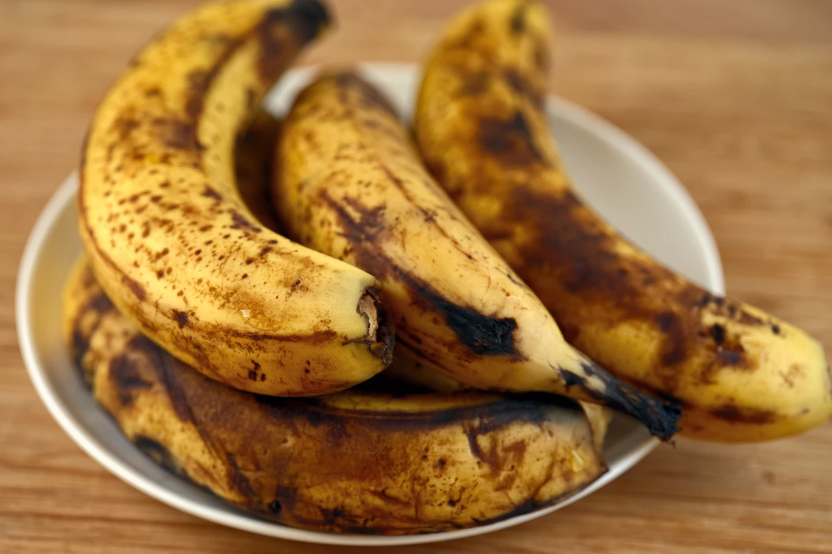 Un manojo de plátanos demasiado maduros con manchas marrones que descansan sobre un plato blanco en una mesa de madera