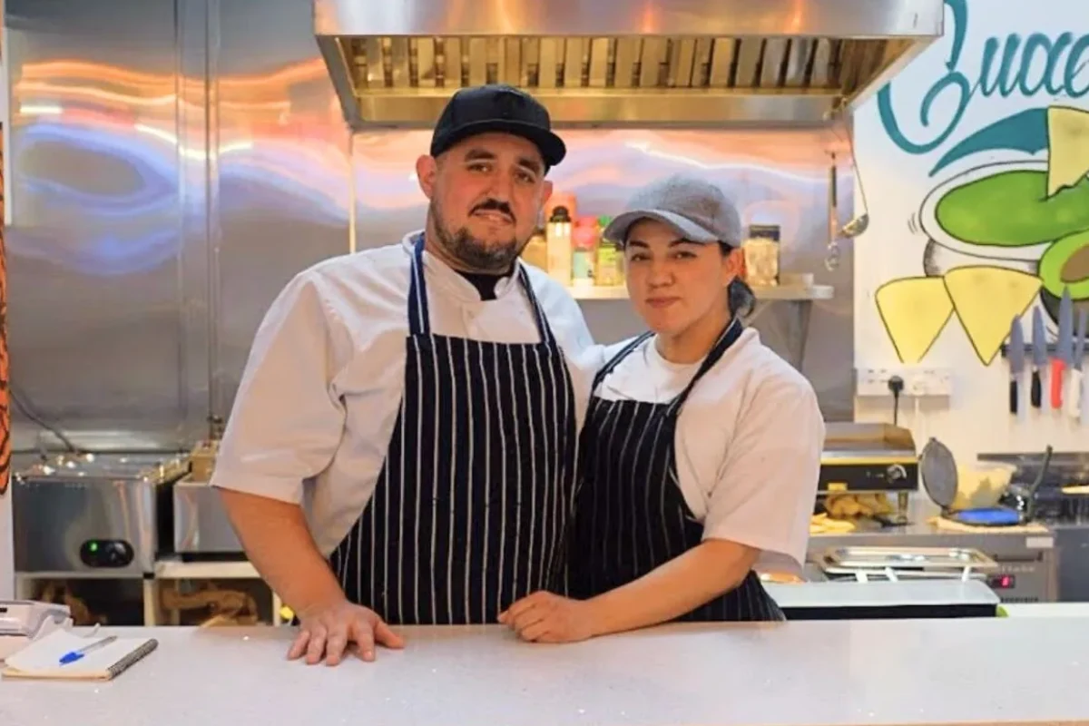 Una pareja de jóvenes con mandiles y gorras sonríen a la cámara al interior de una cocina.