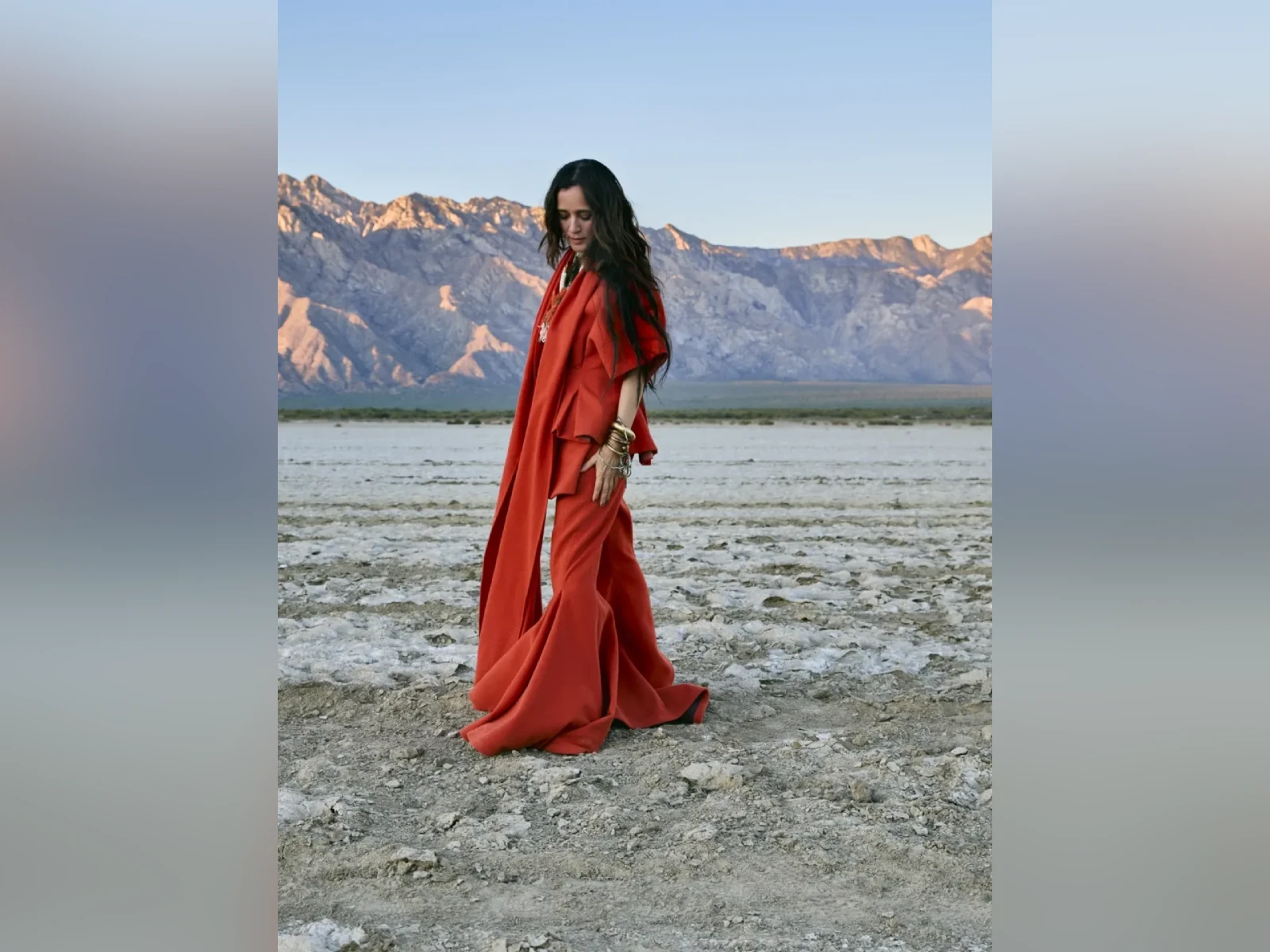 Julieta Venegas en un paisaje desértico con montañas atrás y un vestido rojo-naranja