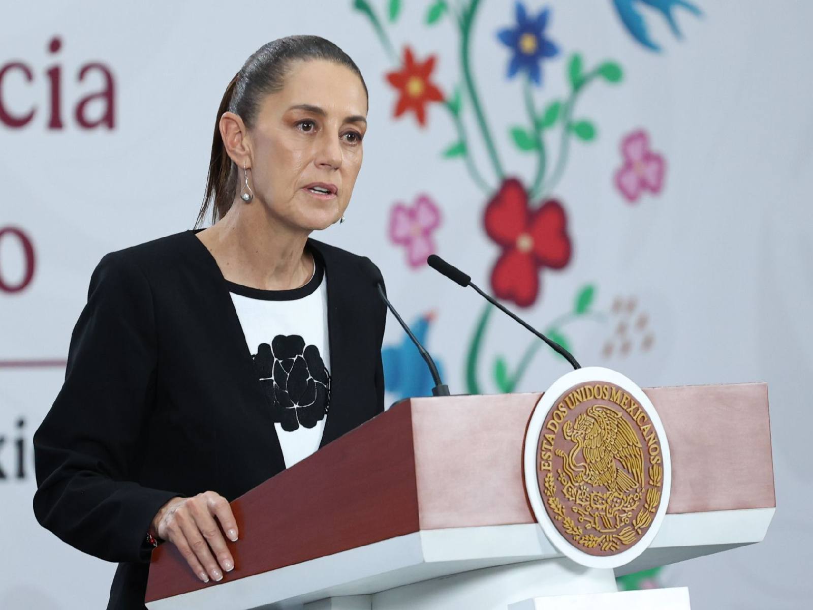 Presidenta Claudia Sheinbaum durante su conferencia de prensa matutina vestida con saco negro y blusa blanca con una flor negra en el centro brodada