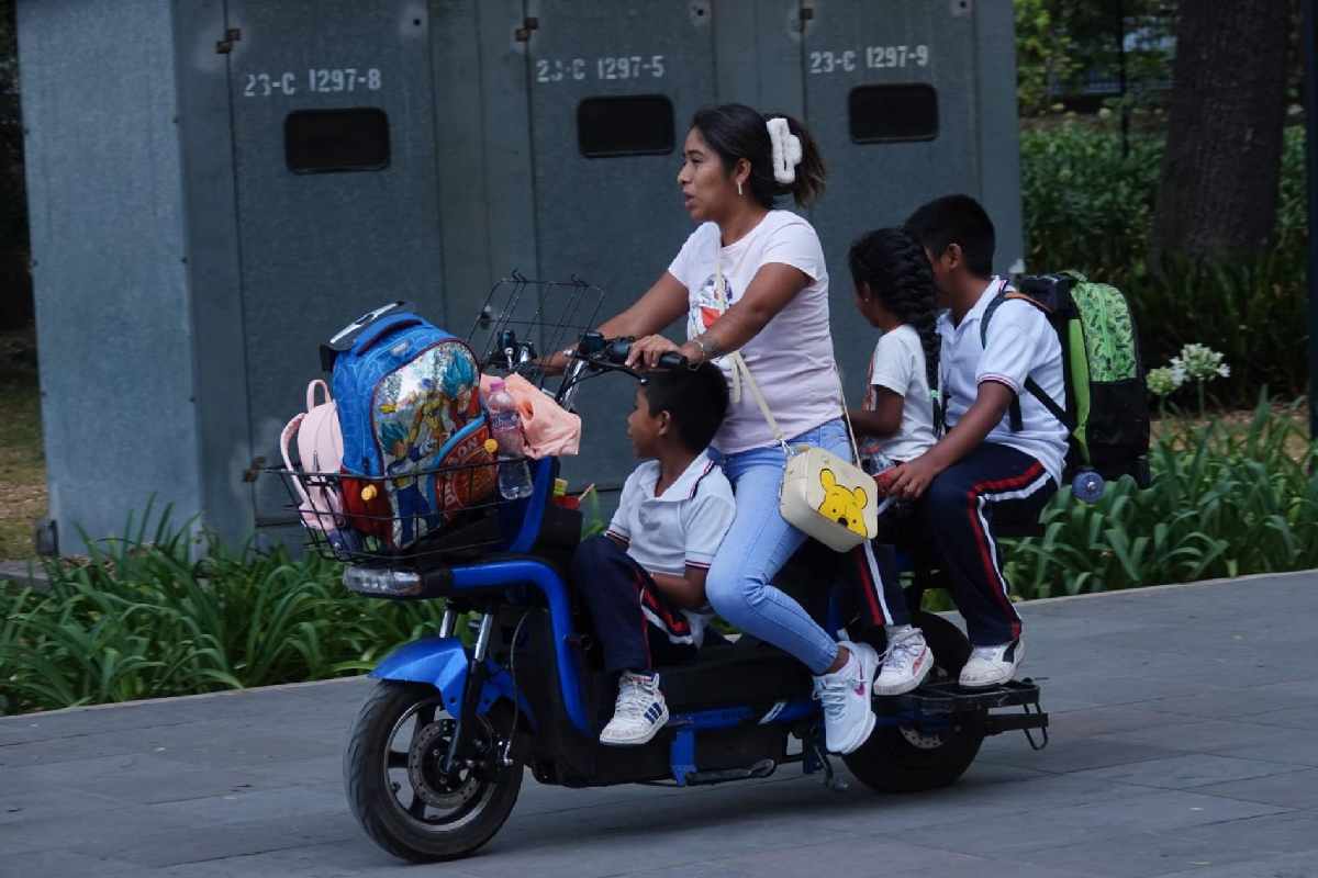 Una mujer viaja con tres menores de edad uniformados todos sobre una bicicleta eléctrica. Al frente hay una pequeña canasta con la mochila de los niños. 