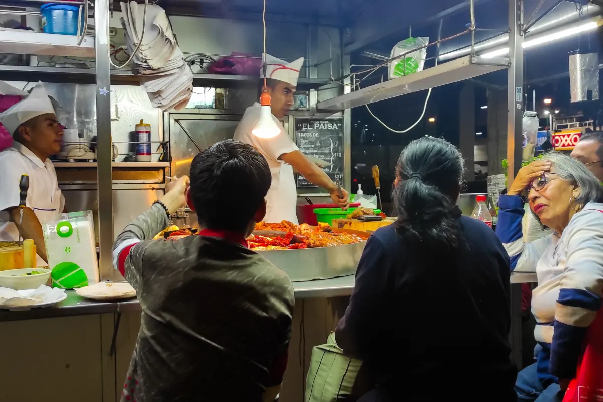 Taquería con gente esperando tacos mientras taqueros se encuentran despachando