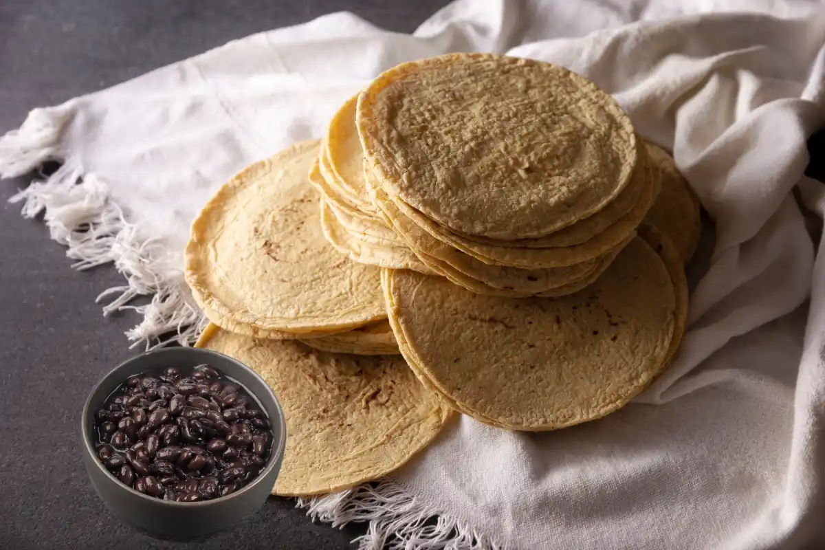 Sobre una mesa está un tela blanco con una torre de tortillas a un costado hay un recipiente con frijoles de la olla