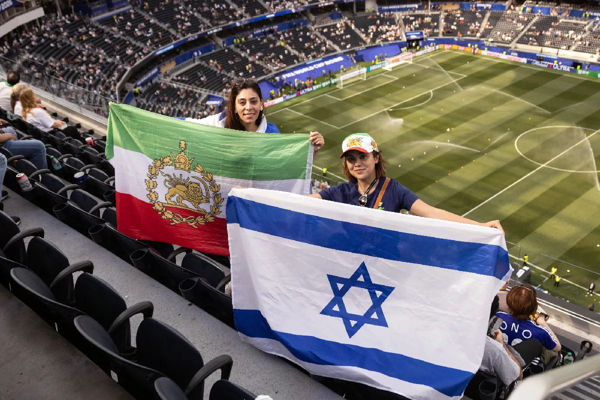 Dos mujeres sosteniendo banderas, una la del sol y el léon; otra, la de Israel. Ambas se encuentran en las gradas del estadio donde jugó IrÁN