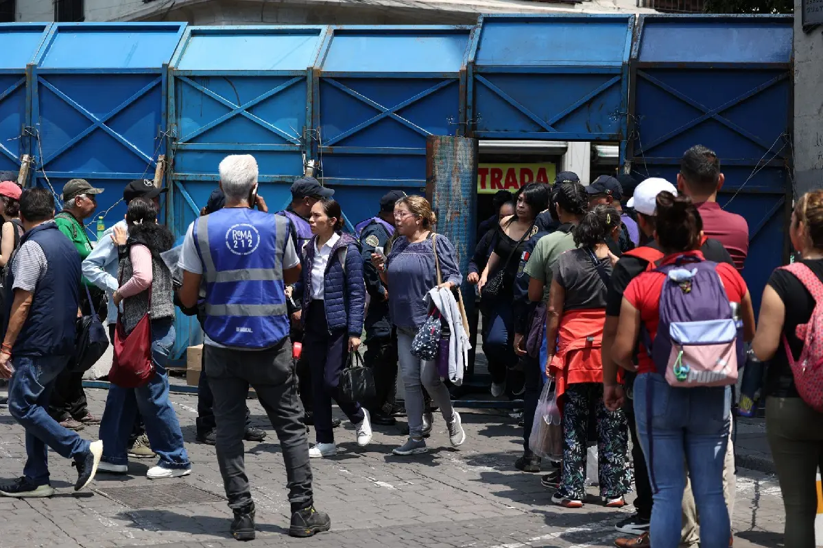 Personas hacen fila para ingresar por una puerta de una valla azul 