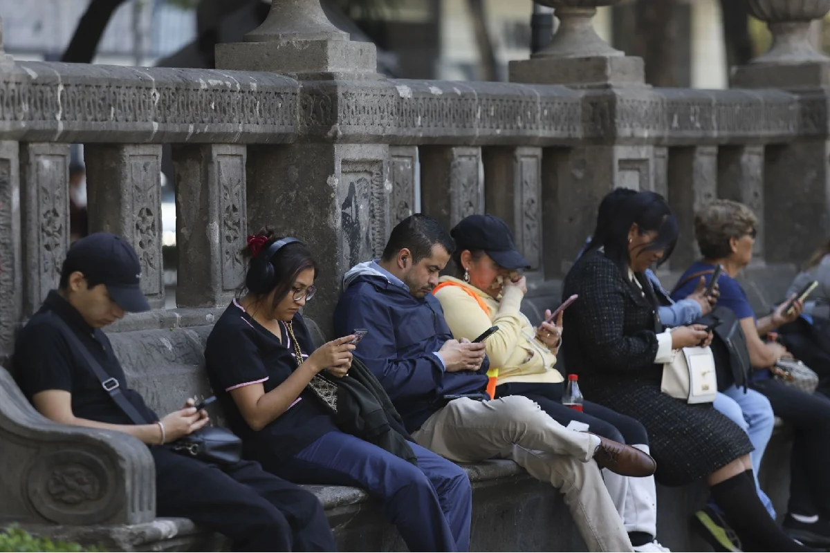 Un grupo de personas sentadas usando sus celulares en vía pública. 