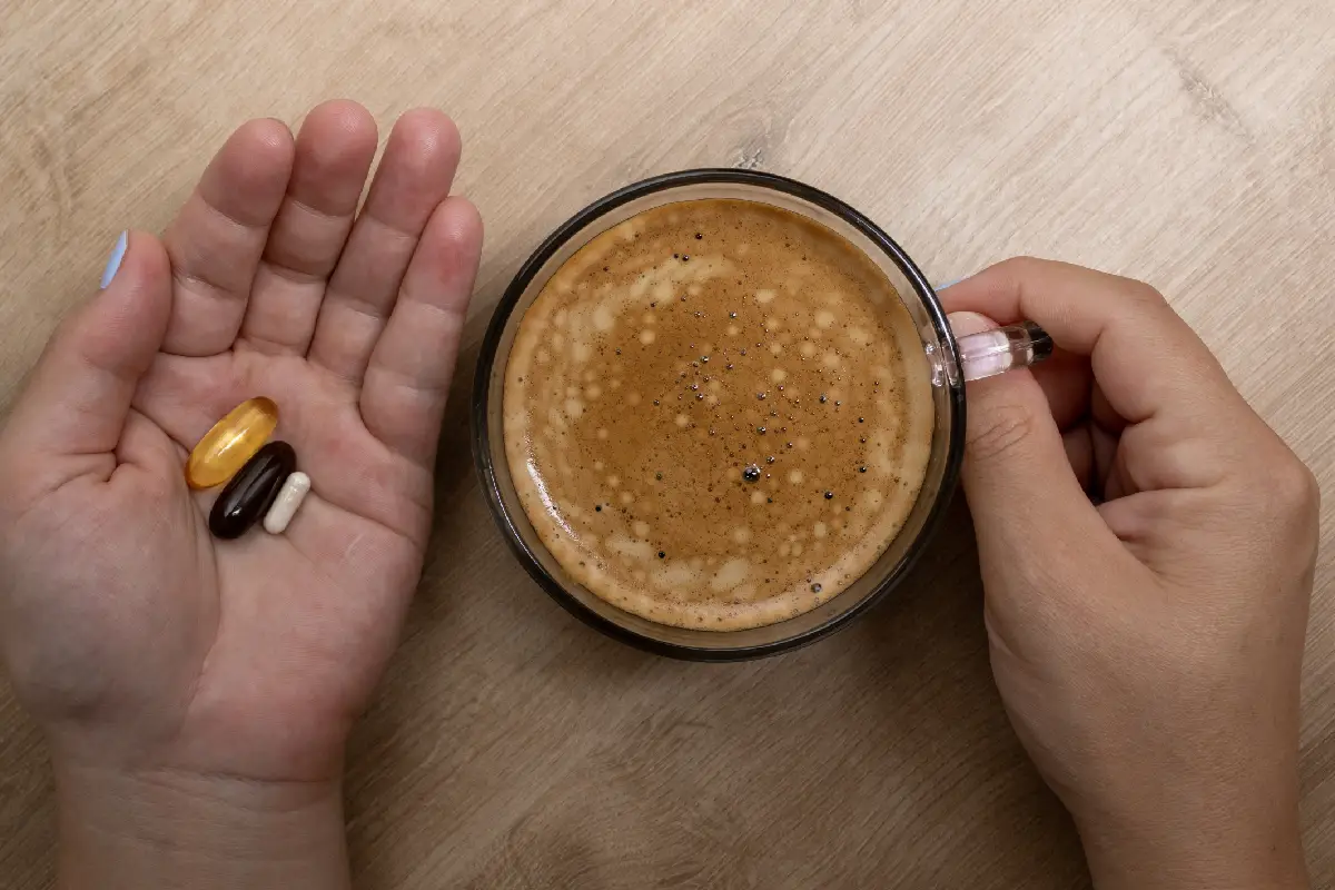 Una manos sostiene pastillas y la otra una taza de vidrio con café