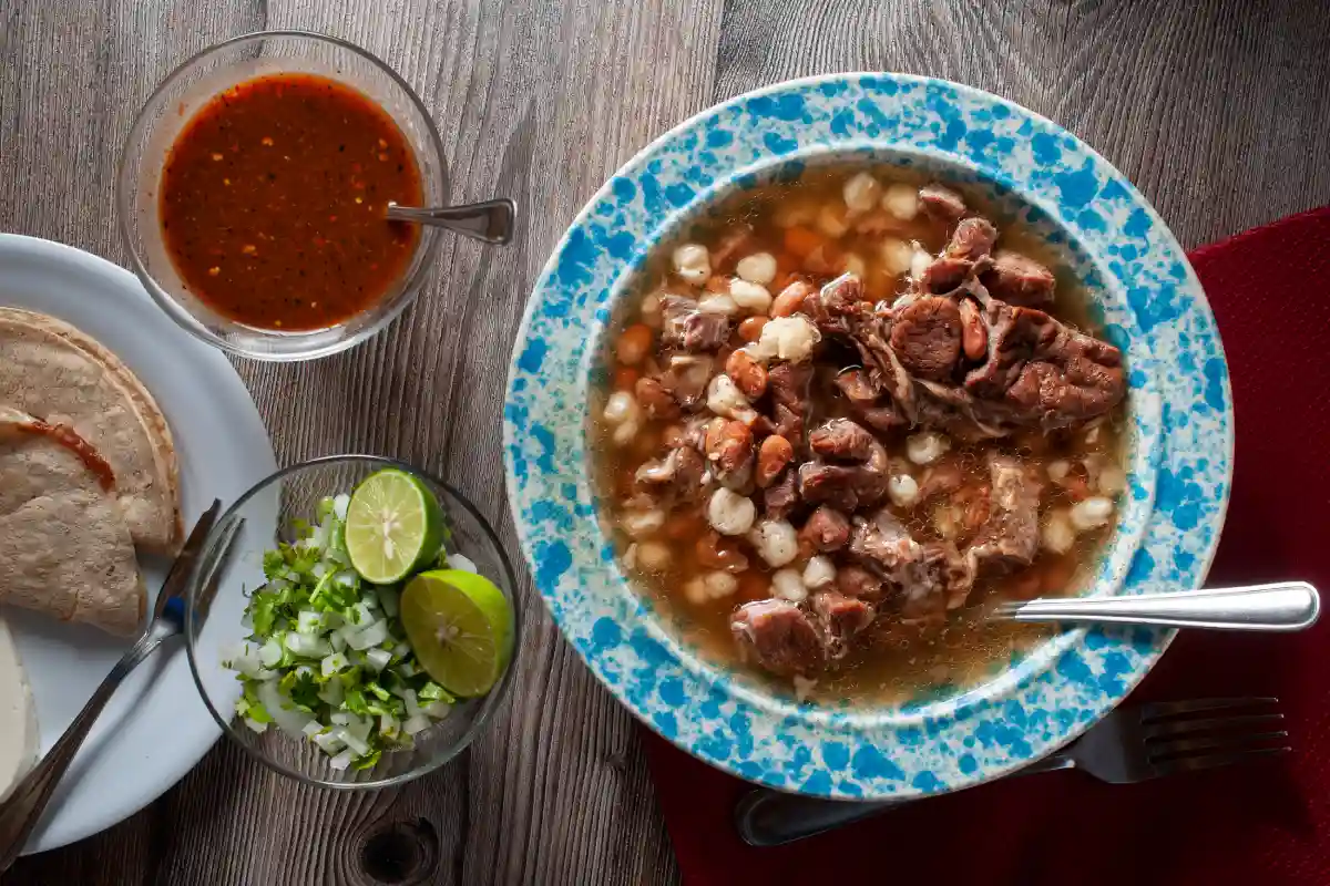 Plato con un caldo con carne acompañado de cilantro y cebolla picada, junto con salsa roja y tortillas