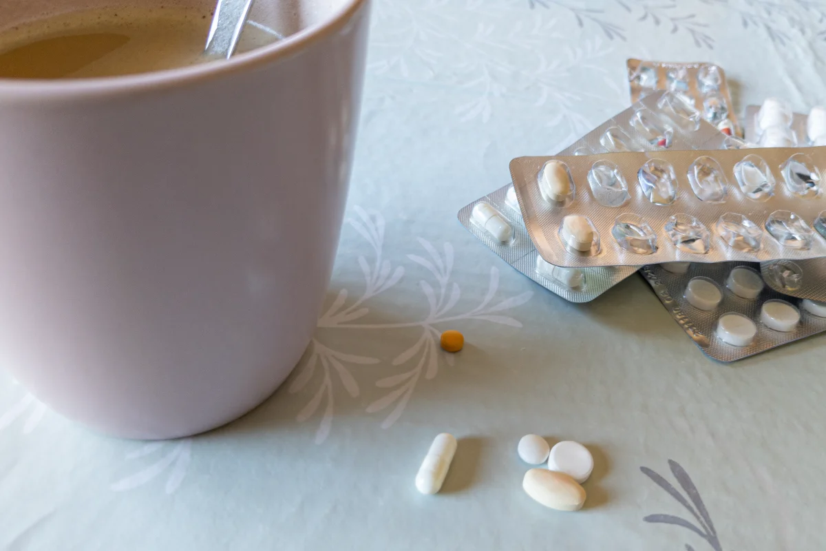 Desayuno con una taza de color rosa claro lleno de café con leche. Lleva una cuchara en el interior para remover la mezcla. En la estera de plástico también hay varias píldoras sueltas de diferentes medicamentos 