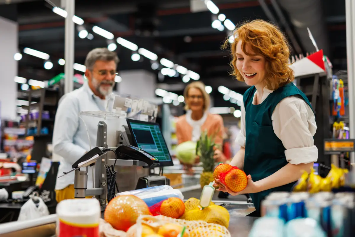 Cajero sonriente que escanea la fruta en el Completar compra de un supermercado con los Clientes que esperan en el fondo.