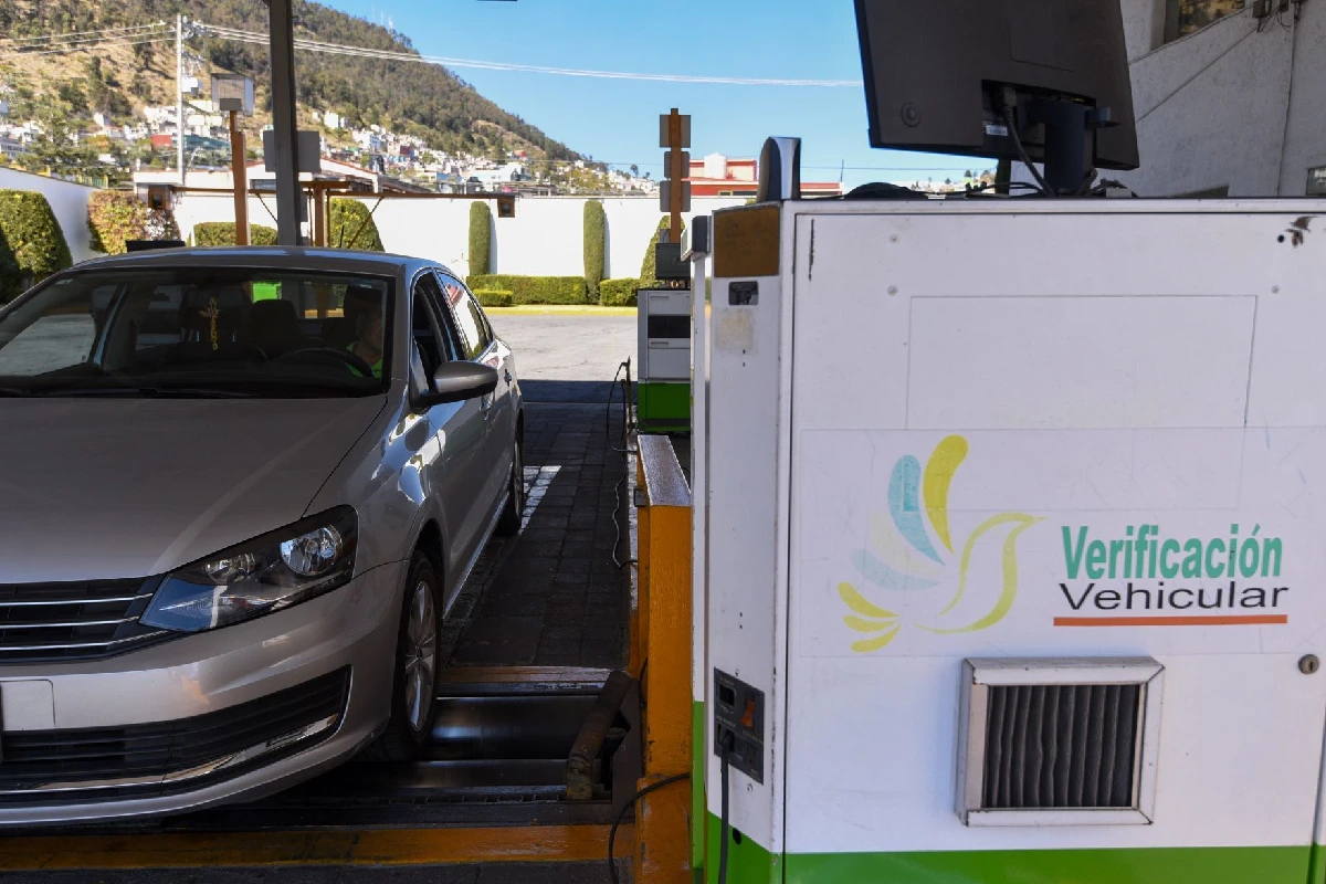 Texto Alt: Primer plano de un módulo oficial de medición en un verificentro del Estado de México, con un logotipo que simula un ave multicolor y la frase "Verificación Vehicular". A la izquierda, un automóvil sedán de color plata se encuentra posicionado sobre los rodillos metálicos de la línea de inspección, listo para la prueba de emisiones obligatoria. Al fondo, se aprecia una zona montañosa con casas bajo un cielo despejado.