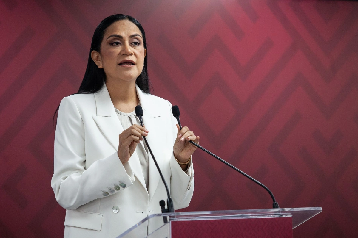 Ariadna Montiel vistiendo de traje blanco y de fondo una pared con grecas color guinda