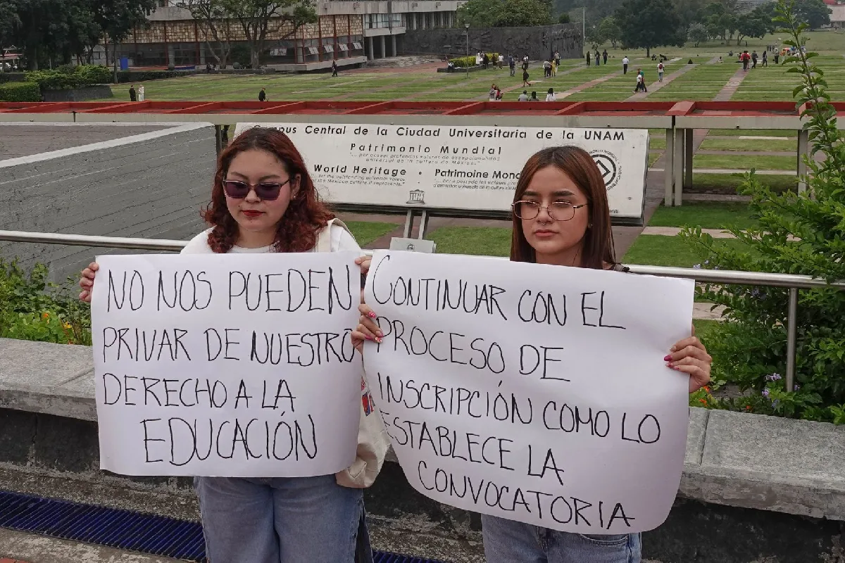 Dos estudiantes protestando frente al letrero de Patrimonio Mundial en Las Islas de la UNAM, sosteniendo pancartas que exigen su derecho a la educación y la continuación del proceso de inscripción según la convocatoria.