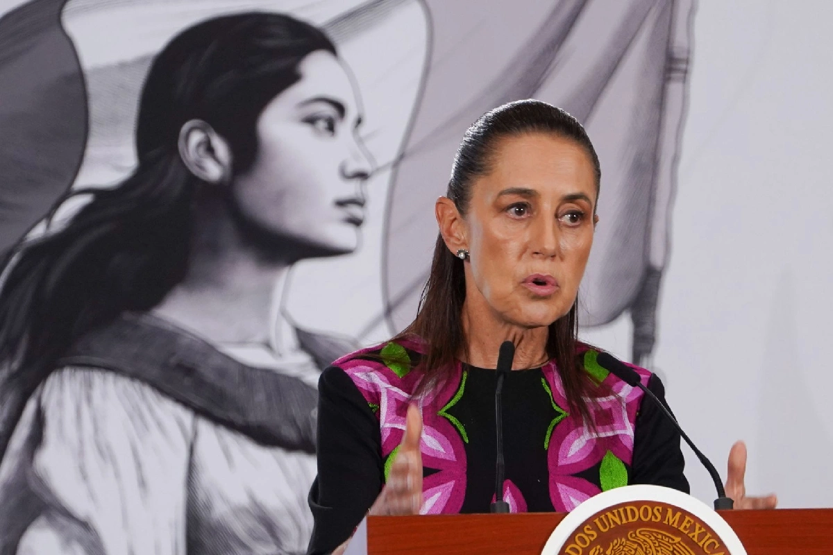 La presidenta Claudia Sheinbaum habla en pódium con las manos extendidas durante una conferencia, con la imagen de Margarita Maza de fondo.