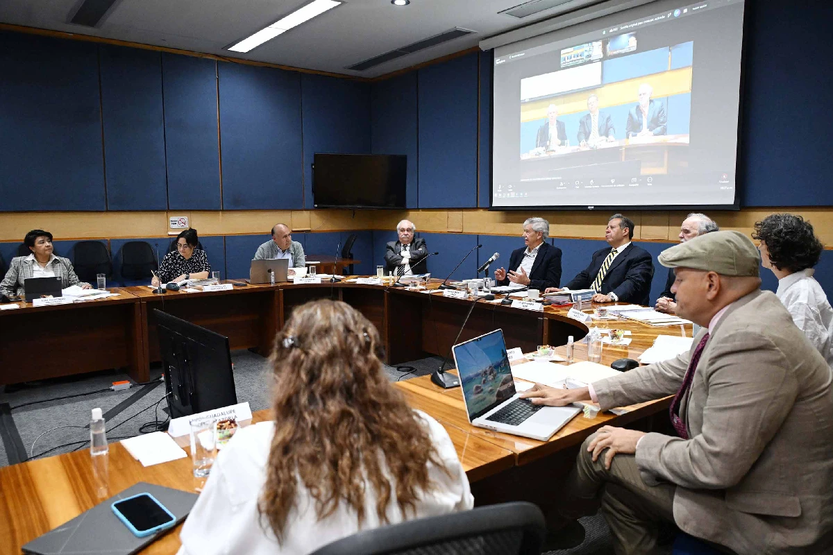 Reunión de la comisíon de expertos académicos sentados en una mesa semicircular en una sala de juntas de la UNAM con proyección en pantalla gigante.