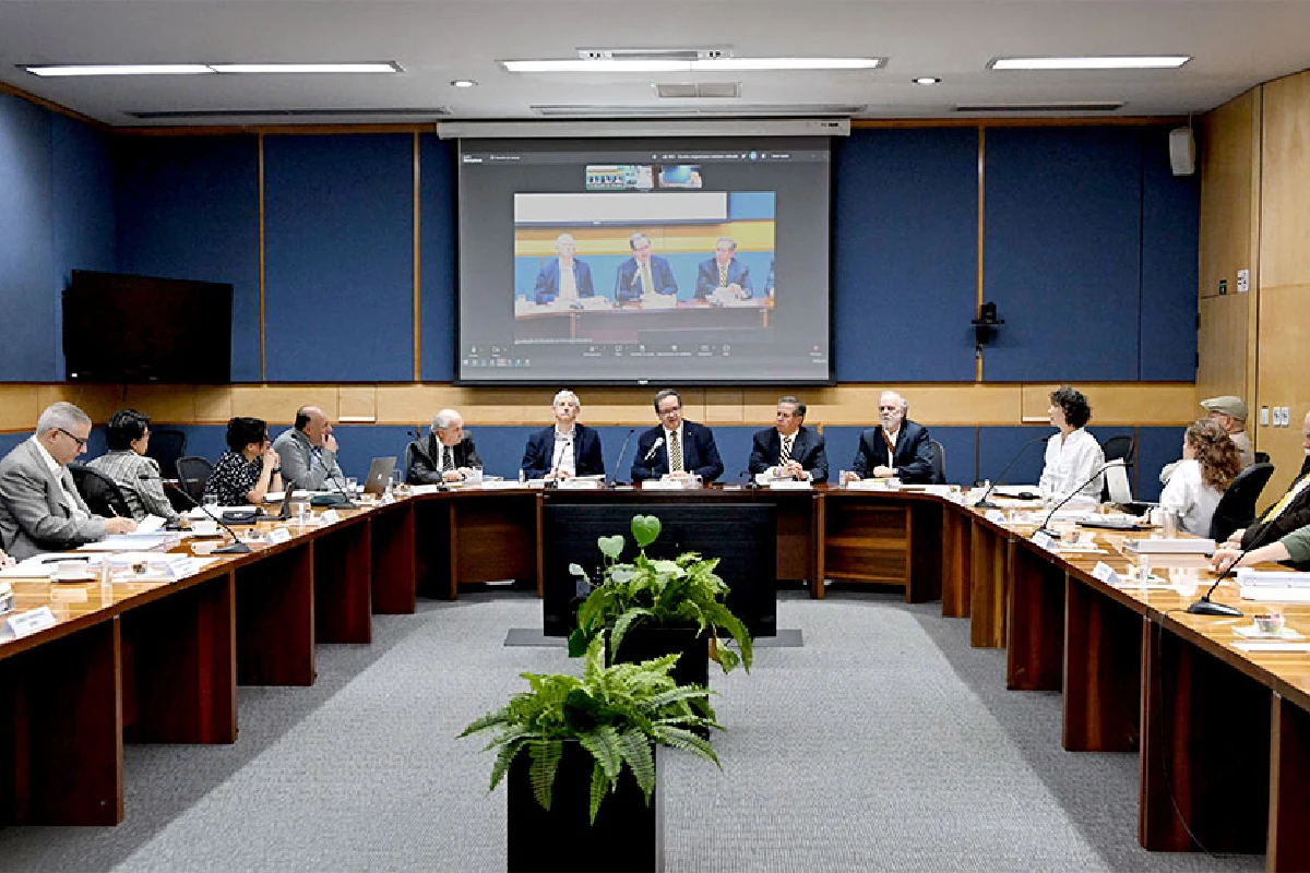 Sesión de consejo o comisión académica de la UNAM en una sala de juntas semicircular, con autoridades universitarias reunidas y una pantalla transmitiendo la reunión por videoconferencia.