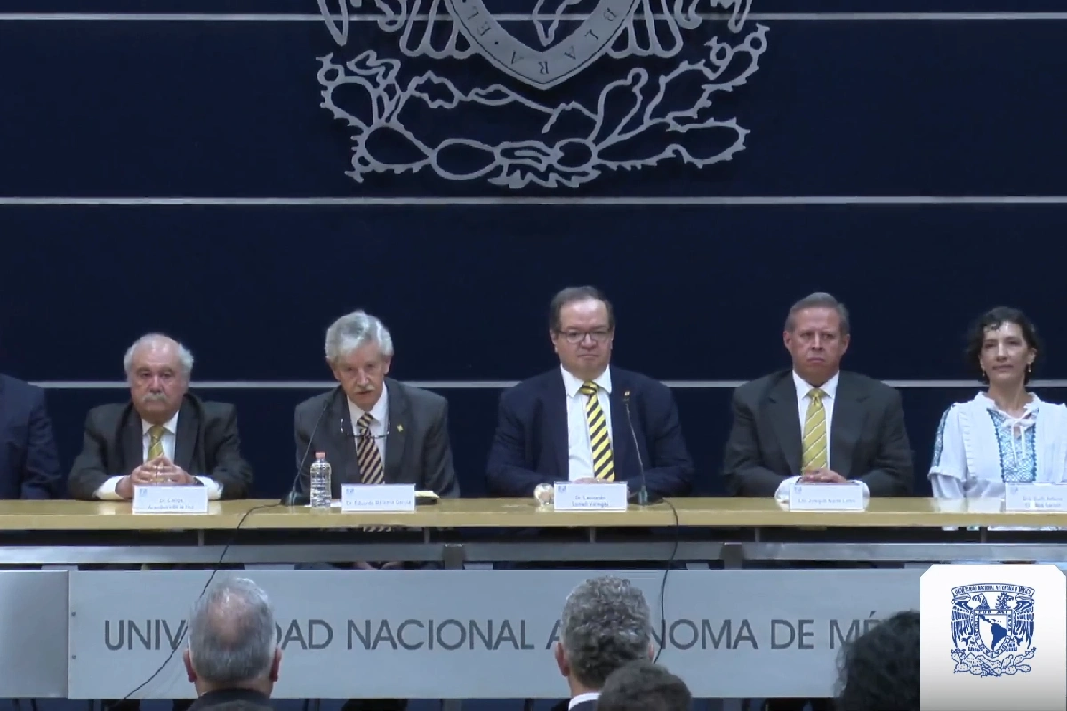 Integrantes de la comisión académica de la UNAM sentados en presídium durante una conferencia de prensa con el escudo institucional de fondo.