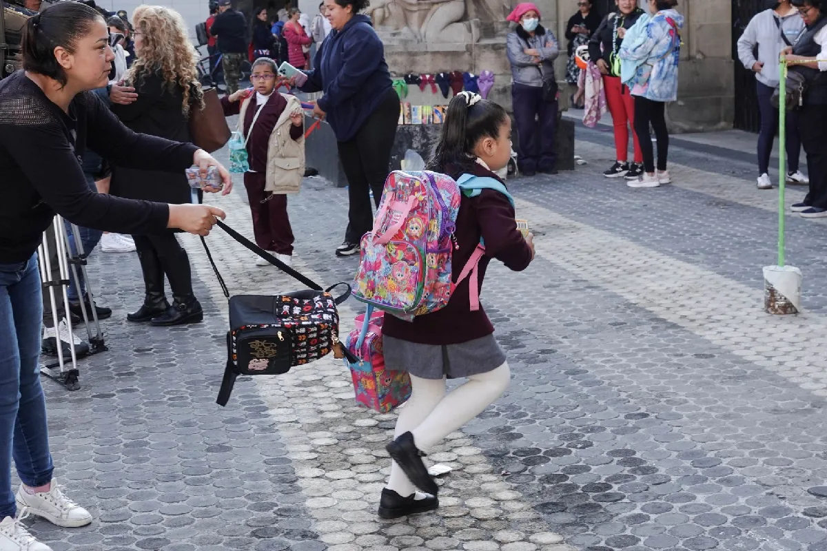 Una niña de primaria con uniforme escolar y mochila de colores camina apresurada hacia la escuela en una calle empedrada mientras una mujer le sostiene la lonchera.