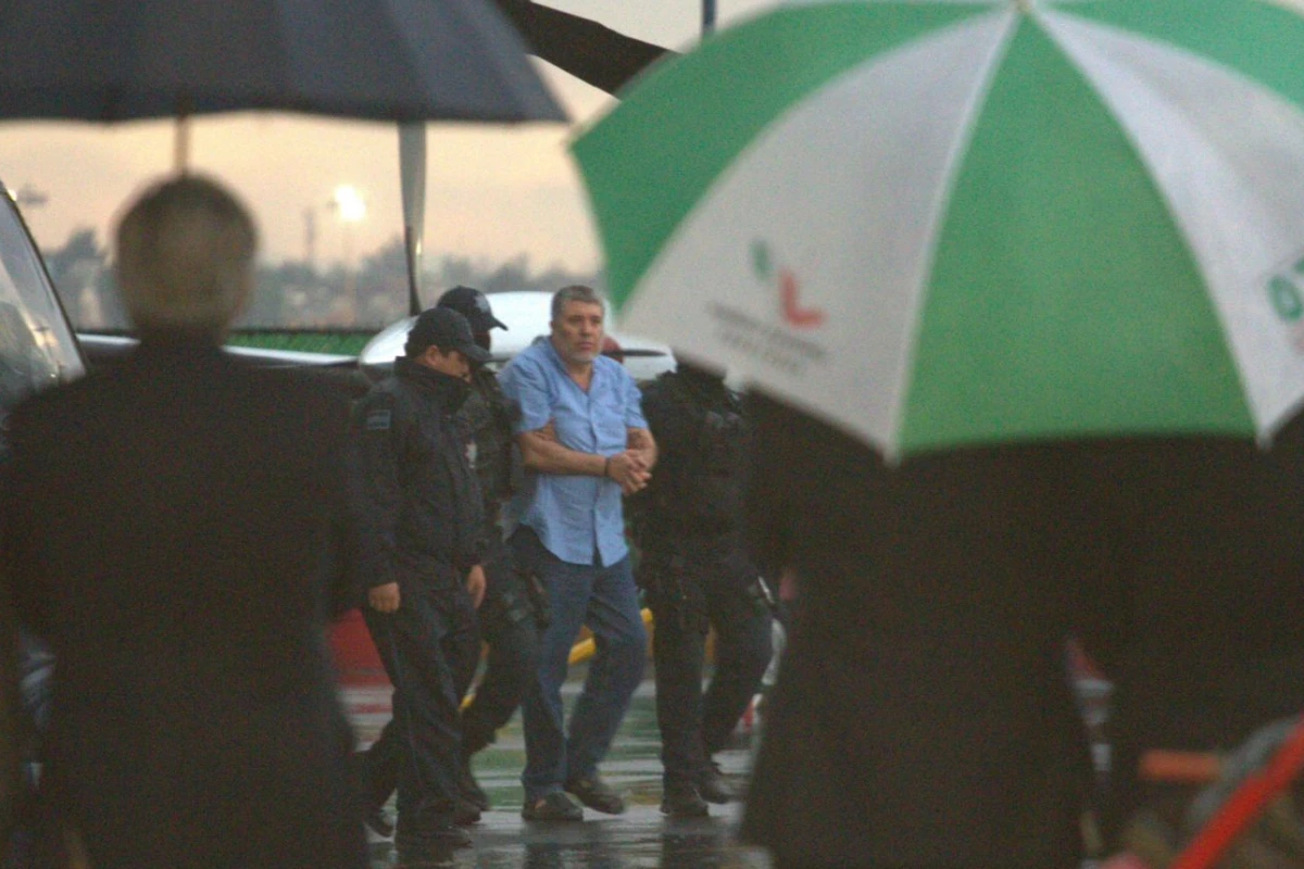 Vicente Carrillo Fuentes, alias "El Viceroy", es custodiado por agentes armados bajo la lluvia en un hangar aeroportuario con helicópteros de fondo.