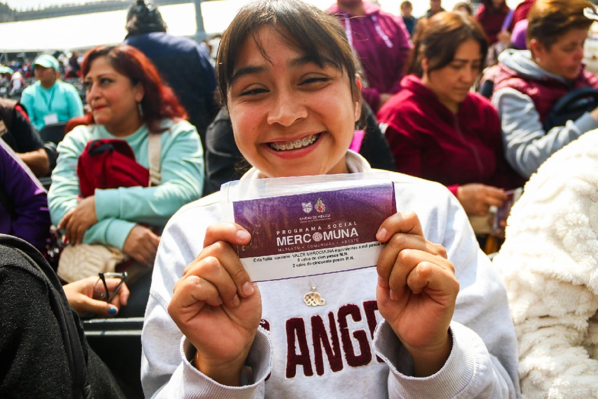 Joven sonriente muestra a la cámara su fajilla de vales del programa social Mercomuna durante un evento masivo de entrega en la Ciudad de México.