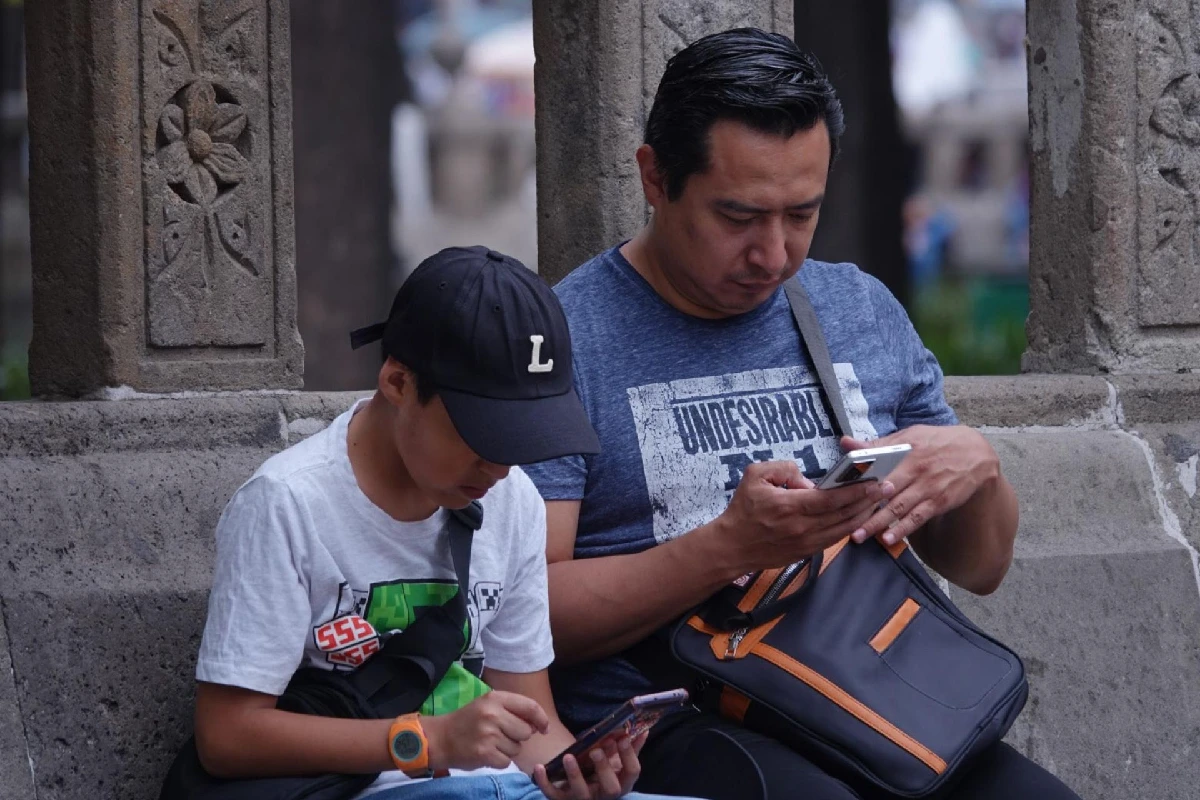 Un niño observando su celular junto a un adulto que también observa su celular.