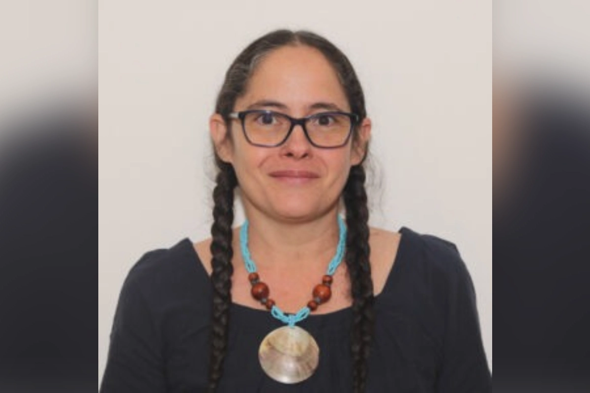 Retrato de la antropóloga cubana Hilda Landrove, sonriendo con gafas de armazón oscuro, peinado de dos trenzas y un collar artesanal de cuentas azules con un dijes circular.