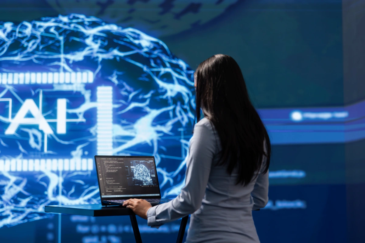 Mujer de espaldas utilizando una computadora portátil sobre una mesa flotante, con una proyección azul de fondo que muestra redes neuronales y la sigla AI.