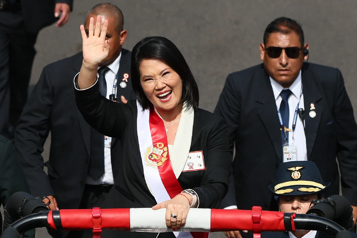 Keiko Fujimori sonríe y saluda con la mano levantada desde un vehículo, portando la banda presidencial de Perú con el escudo nacional.