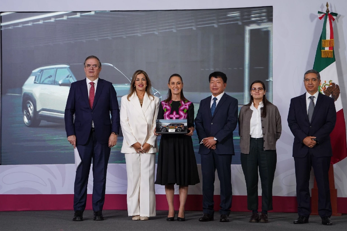 La presidenta Claudia Sheinbaum posa junto a Marcelo Ebrard, directivos de Kia y funcionarios frente a una pantalla con un auto eléctrico, sosteniendo la maqueta de la camioneta de la marca.