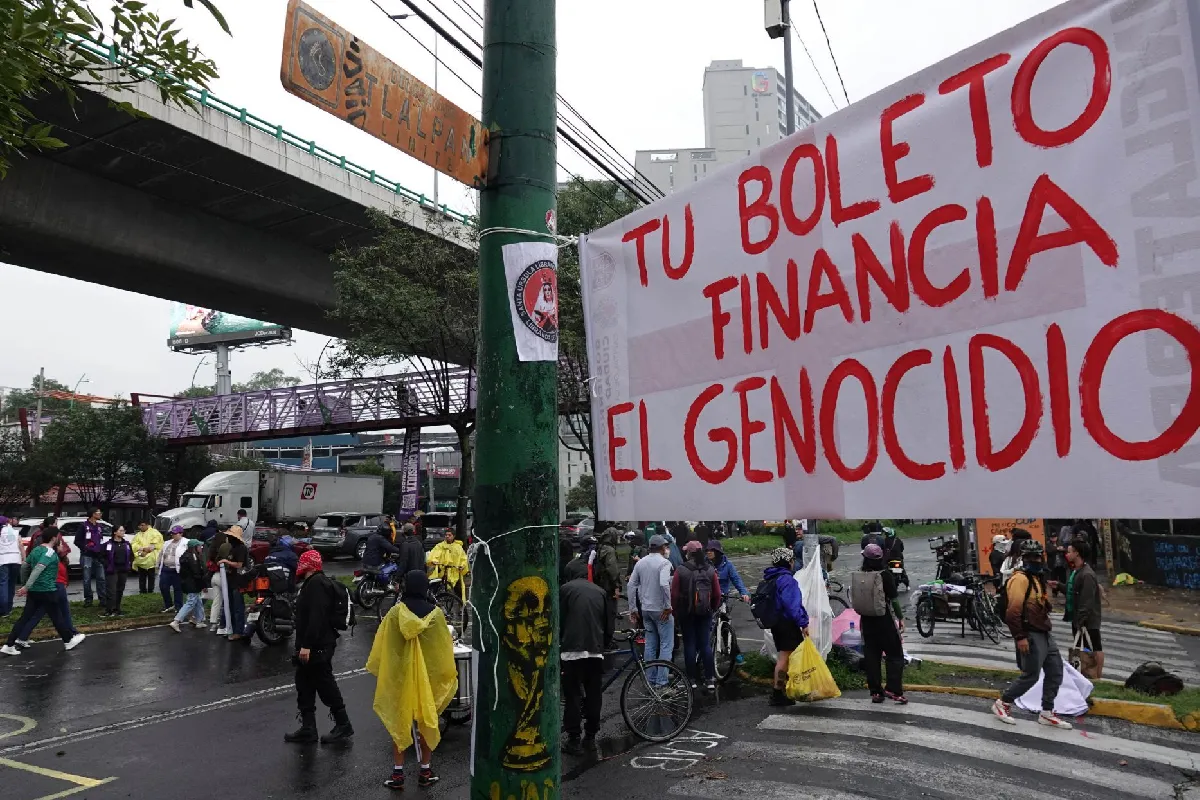 Manifestantes bloquean vialidad y en un posta hay colgado un letrero que dice "tu boleto financia el genocidio" 