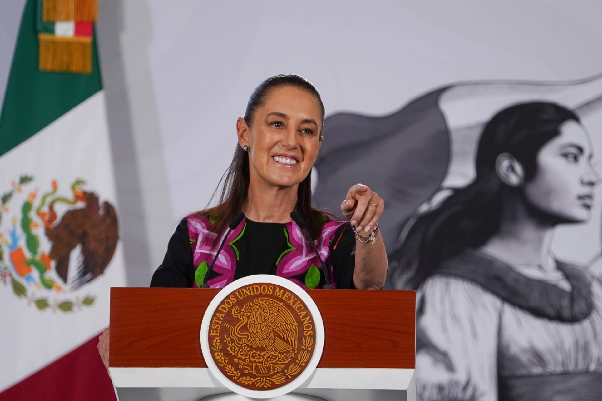 La presidenta Claudia Sheinbaum sonríe y señala hacia al frente desde el pódium oficial con el escudo de México, flanqueada por la bandera nacional.