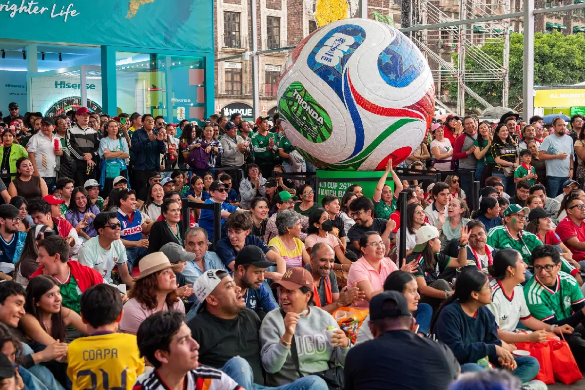 Gran multitud de aficionados vestidos con camisetas de fútbol reunidos en una plaza pública junto a un balón gigante con la marca "TRIONDA" de la Copa Mundial 2026.