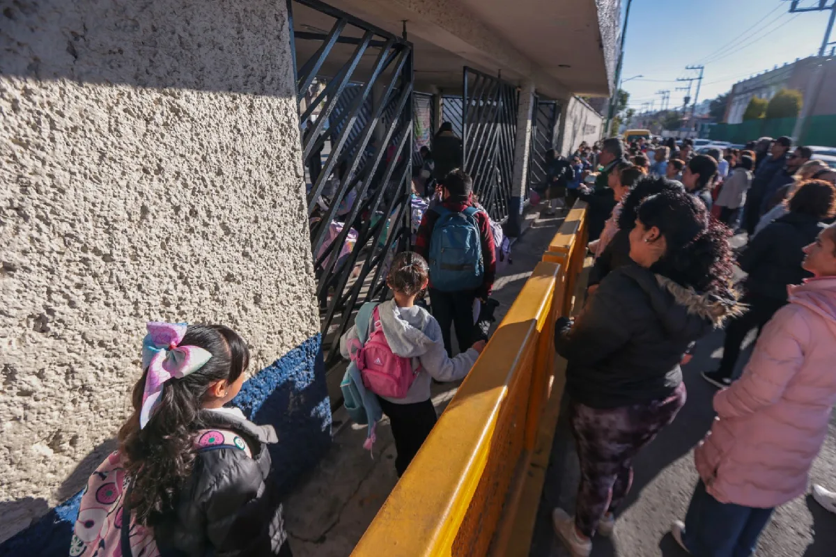 Estudiantes cargando sus mochilas forman una fila para entrar por la reja metálica de una escuela pública, acompañados por sus padres en el exterior.