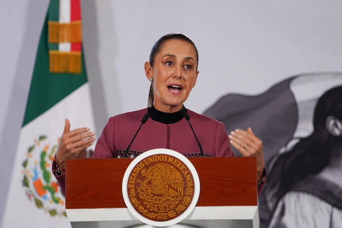 La presidenta Claudia Sheinbaum hablando desde un pódium de madera con el escudo nacional de México y la bandera de fondo durante una conferencia de prensa.