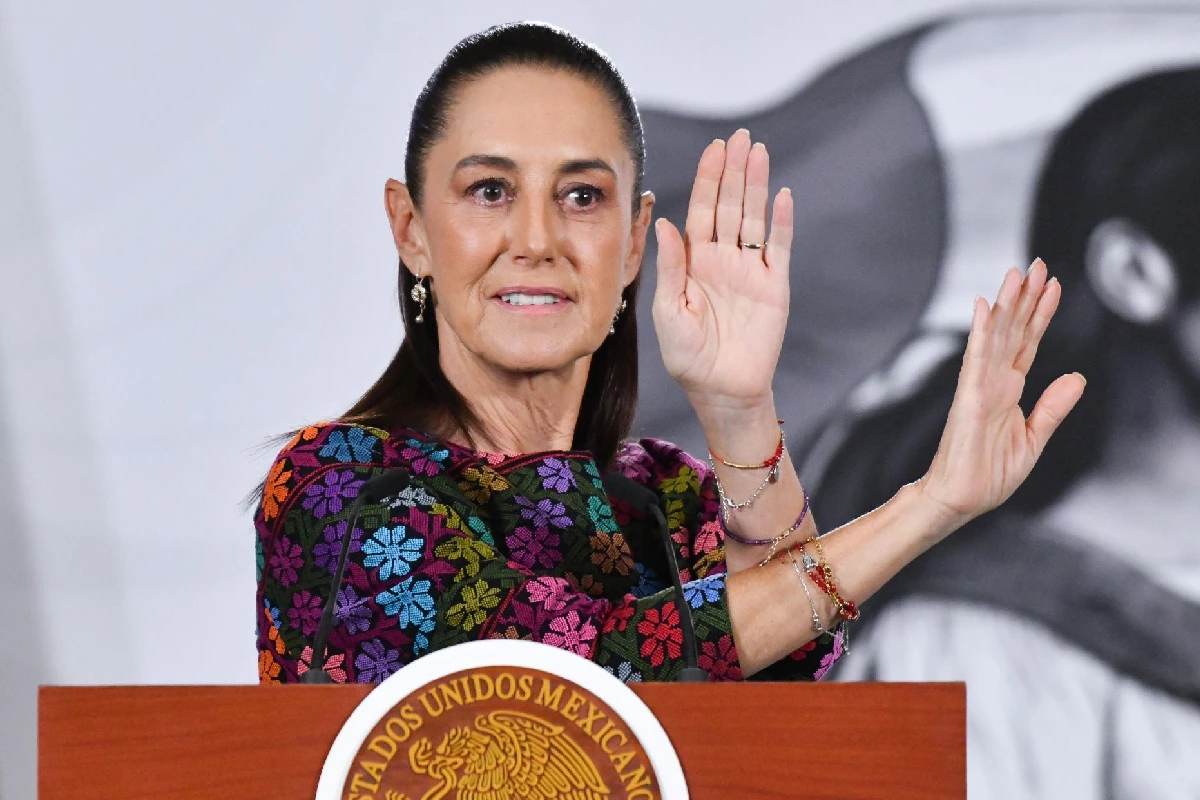 Presidenta Claudia Sheinbaum usando un vestido negro con toques bordados de color sonriendo mientras cruza sus manos al explicar algo 
