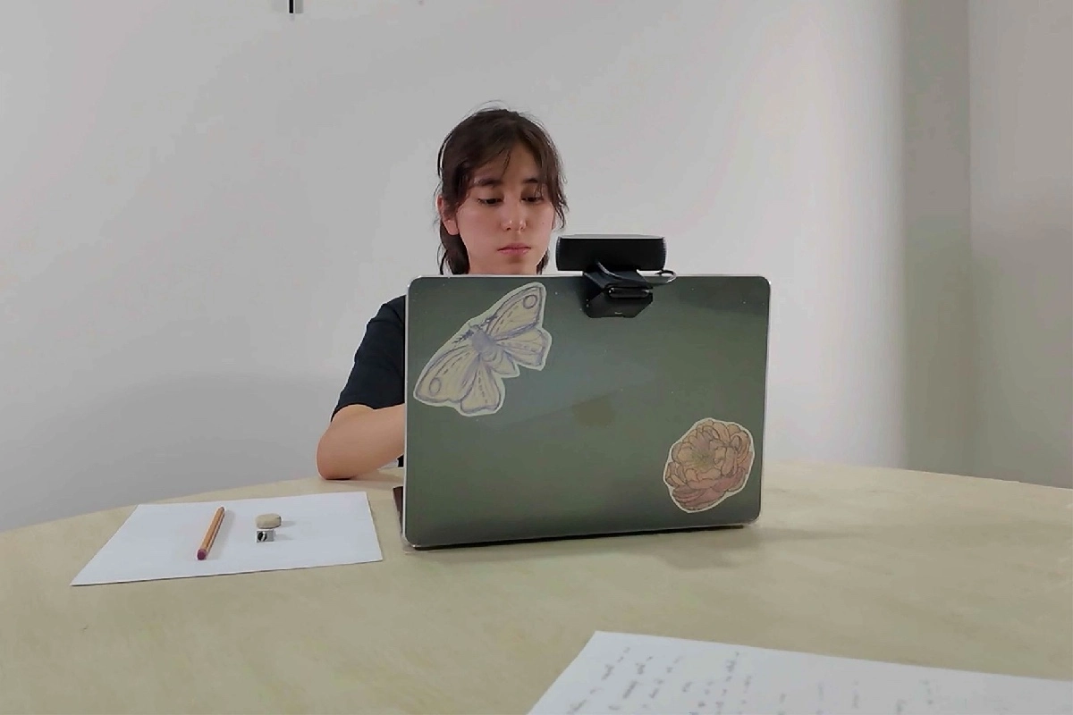 Una joven realiznado su examen en línea en una laptop con cámara y una hoja a su derecha 