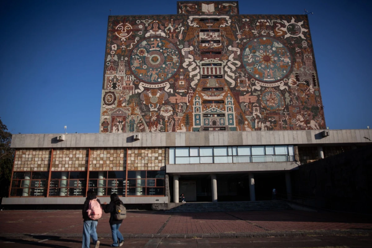 Vista frontal del edificio de la Biblioteca Central de la UNAM con su mural histórico y dos estudiantes caminando por la explanada.