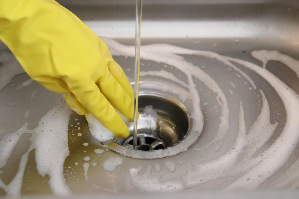 Mano de mujer en guante amarillo sosteniendo una esponja en un fregadero metálico con espuma detergente y agua. 