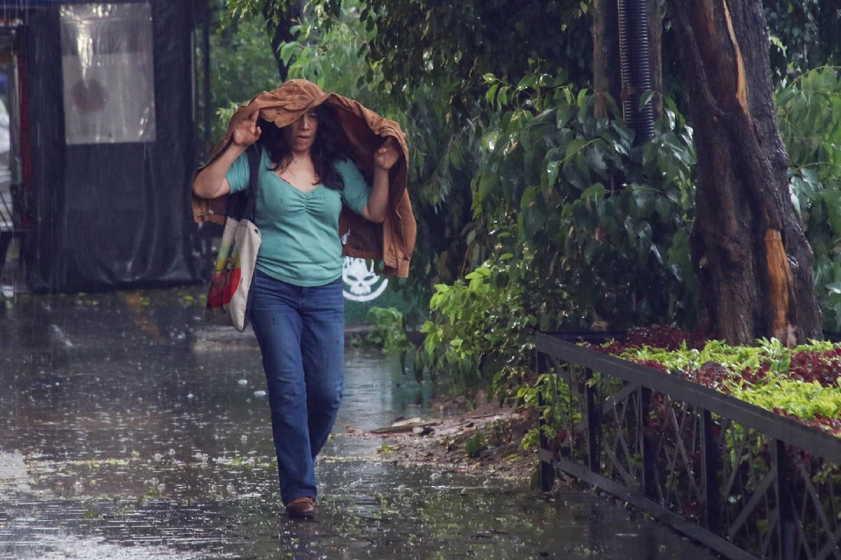 Mujer cubriéndose de la fuerte lluvia con una chamarra sobre la cabeza mientras camina por la banqueta.