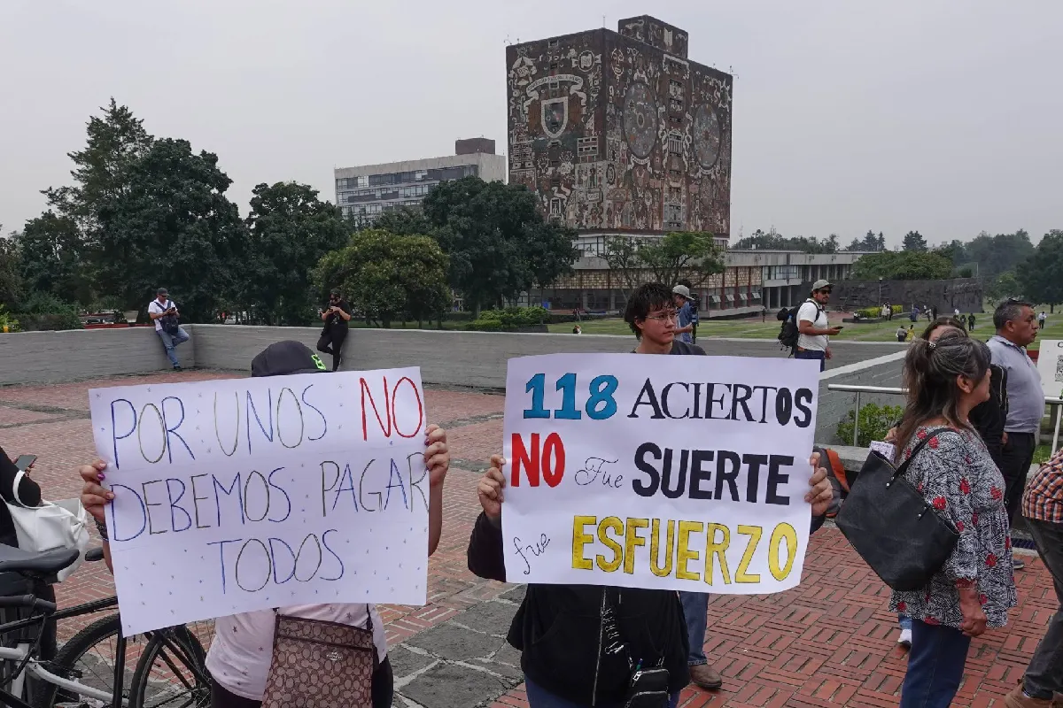 Personas protestando en la explanada de la UNAM con carteles que dicen "Por unos no debemos pagar todos" y "118 aciertos no fue suerte, fue esfuerzo", frente a la Biblioteca Central.