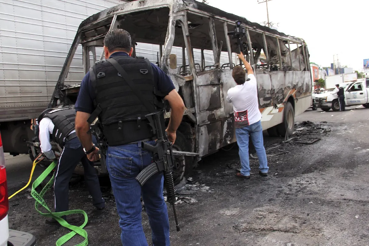 Agente armado observando un autobús de transporte público totalmente incendiado en la calle, mientras autoridades preparan el remolque y un camarógrafo graba la escena.