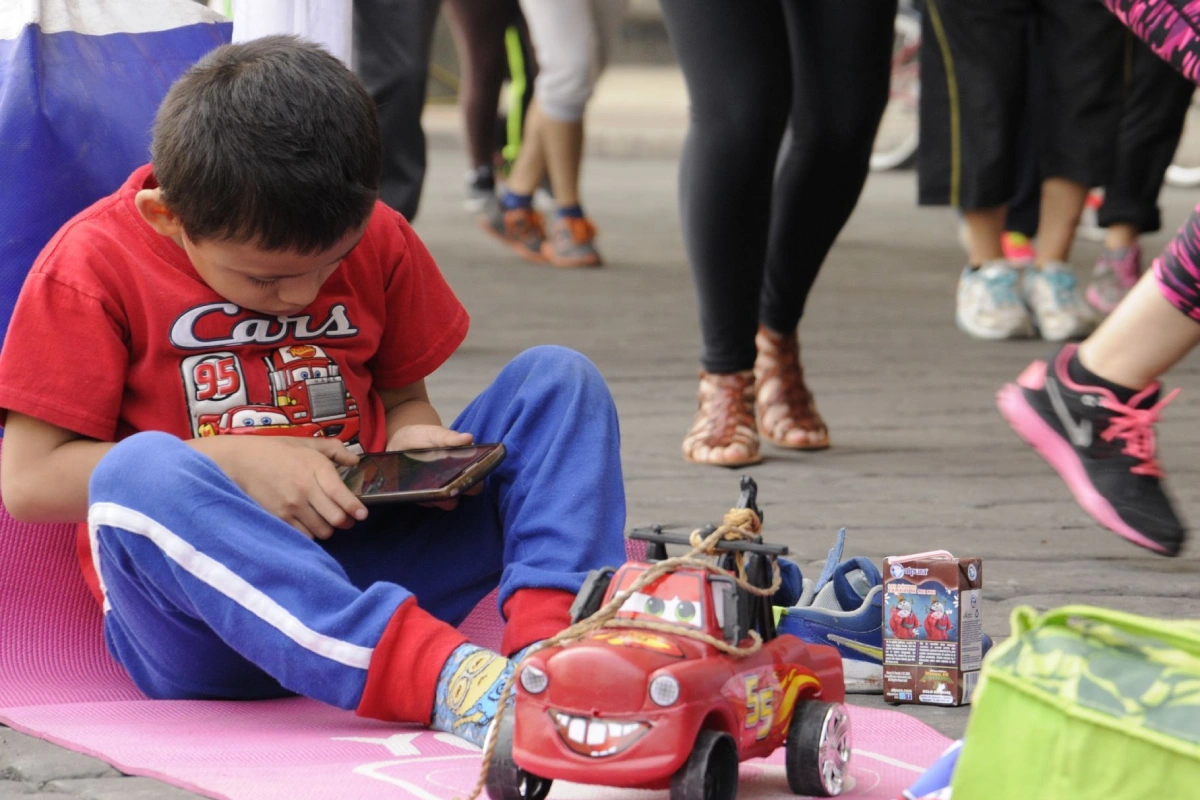 Un niño viendo el celular con sus jueguetes frente a él