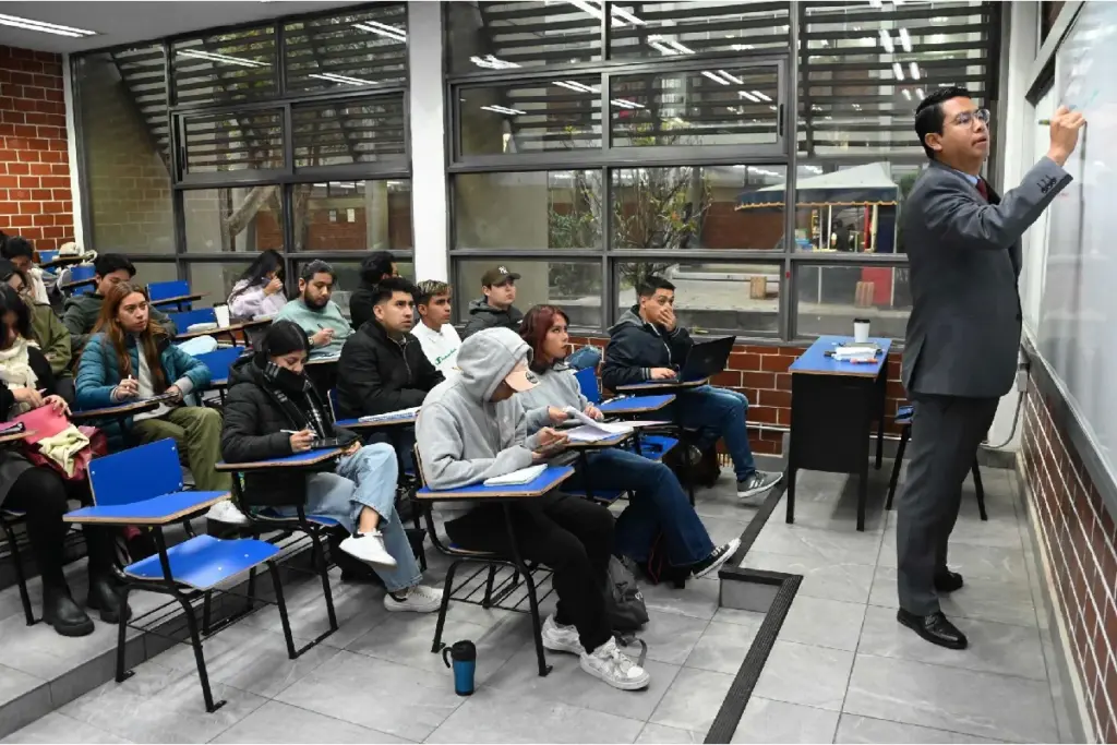 Un profesor en traje escribe sobre un pizarrón blanco frente a un grupo de estudiantes universitarios atentos en un salón de clases.