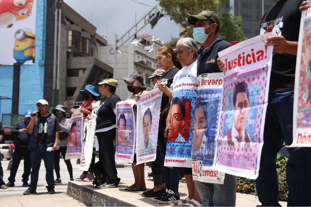 Familiares de los estudiantes desaparecidos de Ayotzinapa parados en fila sosteniendo carteles con rostros y mensajes de "¡Justicia!" y "¡Vivo se lo llevaron!".