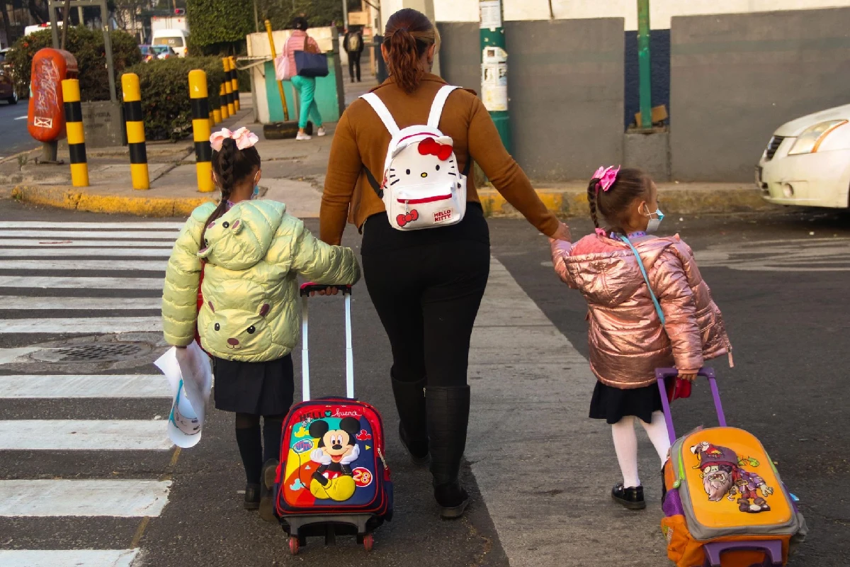 Madre caminando de espaldas junto a sus dos hijas uniformadas arrastrando mochilas con ruedas al cruzar un paso peatonal en el regreso a clases de primaria.