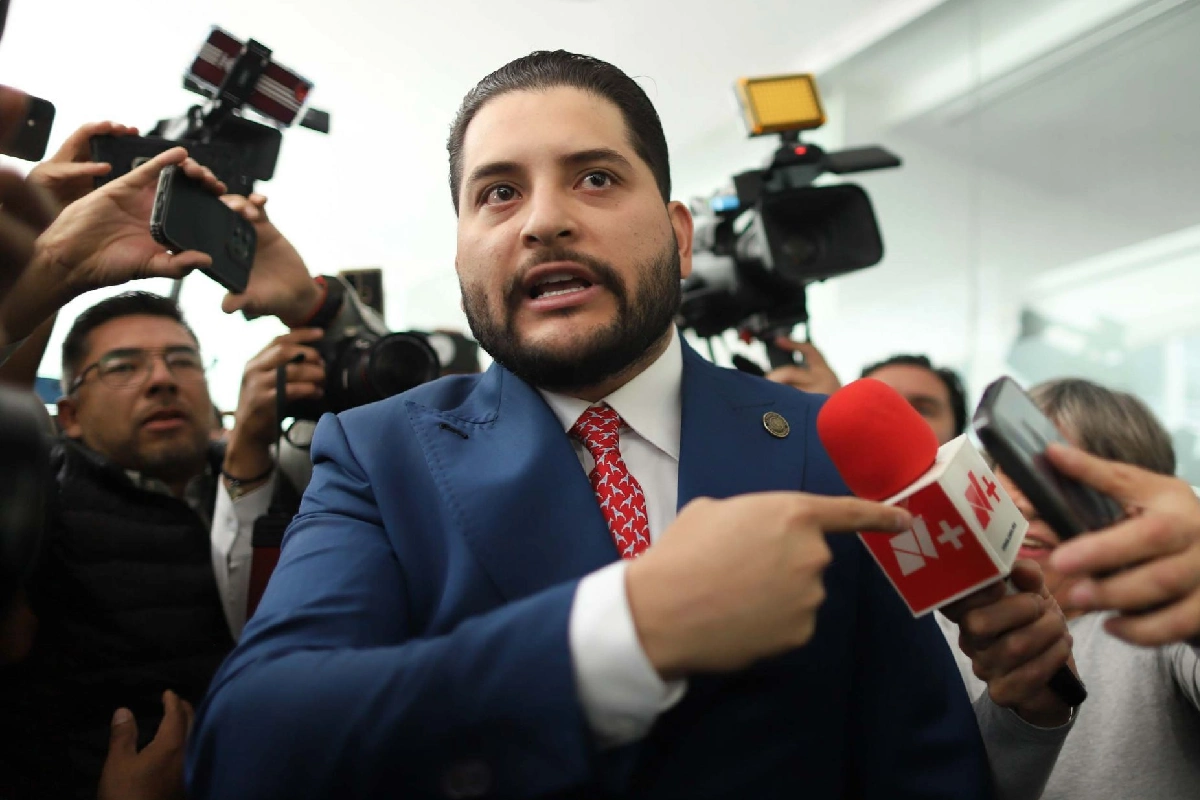 Carlos Gutiérrez Mancilla, con traje azul y corbata roja, gesticulando y señalando hacia un micrófono mientras es rodeado por reporteros y camarógrafos.