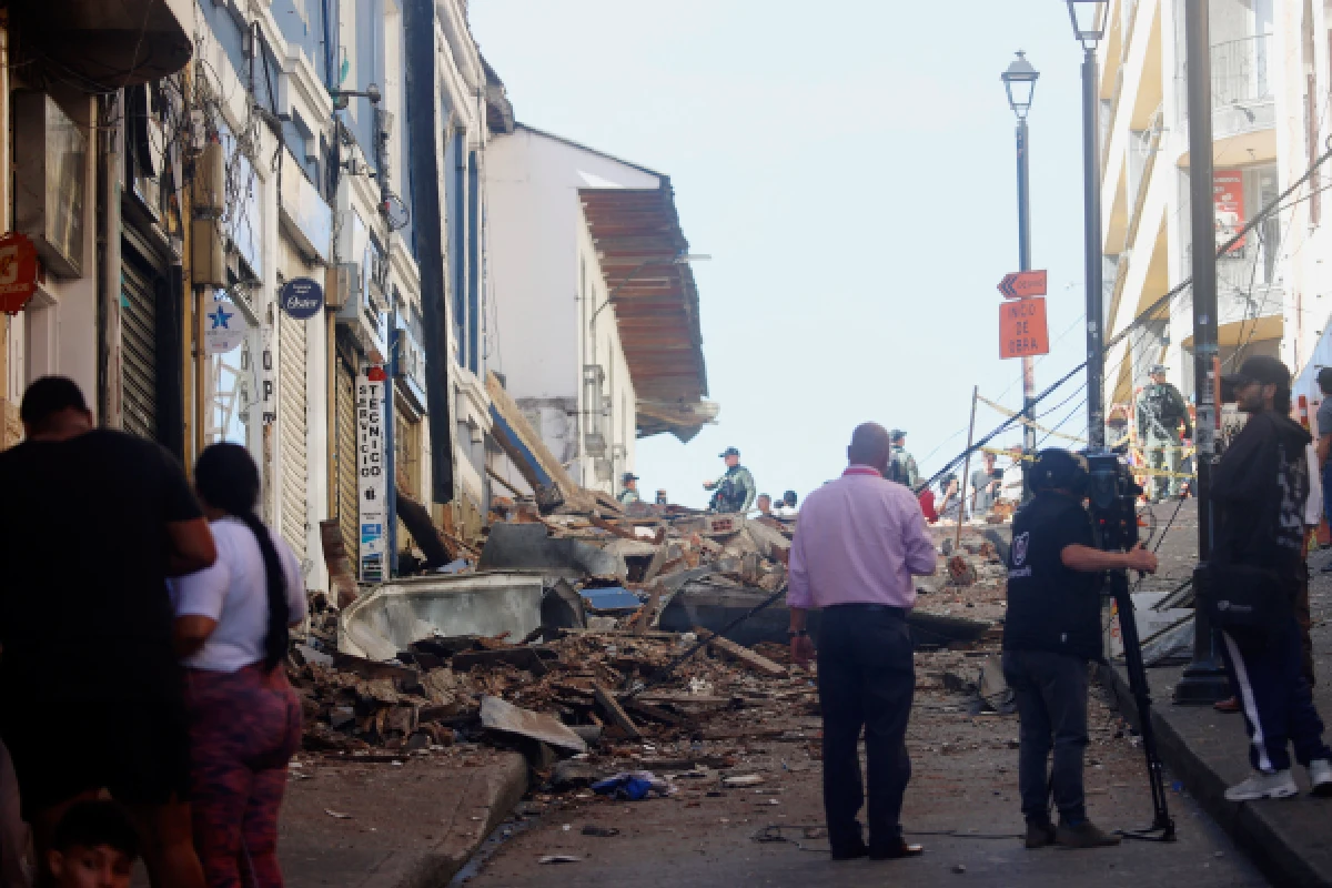 Calle empinada bloqueada por escombros de un derrumbe, con un camarógrafo de televisión documentando los daños y transeúntes observando el lugar.