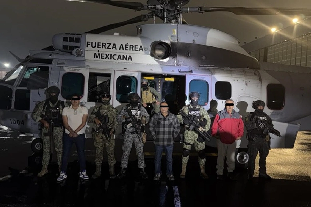 Tres hombres detenidos con la cara censurada posando junto a elementos armados de las Fuerzas Armadas frente a un helicóptero de la Fuerza Aérea Mexicana durante la noche.
